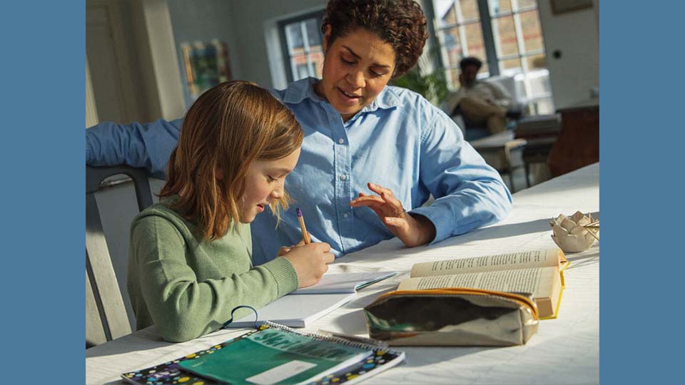 Kvinna sitter vid ett bord med ett barn, de hjälps åt med läxor.