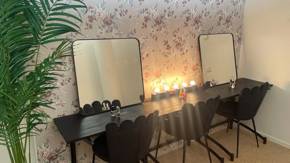 Ett rum med två speglar på ett bord, men tre svarta stolar och en sminklampa. 