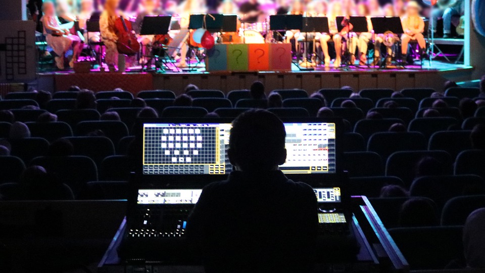 En stor orkester sitter på en scen upplyst i lila och blått ljus, med vimplar över scenen. I förgrunden syns en person bakifrån som sköter ljud och ljus från salongen. Foto