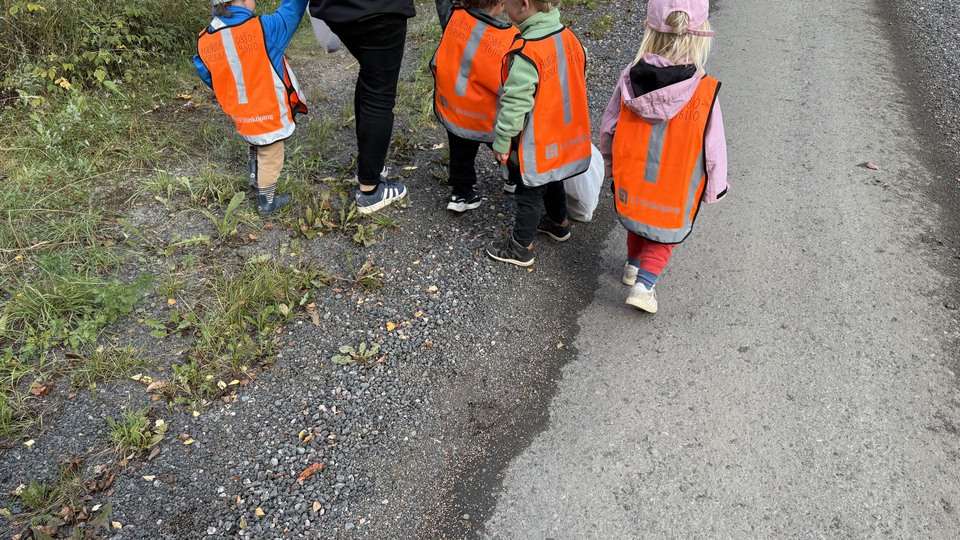 Fyra barn och en vuxen som är ute på promenad och håller i påsar. Barnen har reflexvästar på sig.