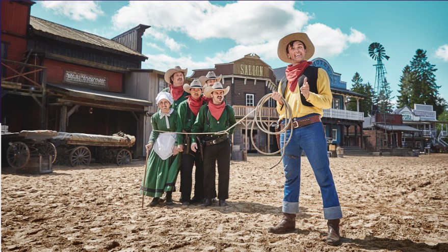 En grupp personer i westernkläder står på en sandig gata i en gammal vilda västern-miljö, med saloon och vindpump i bakgrunden. En person i förgrunden håller ett lasso.