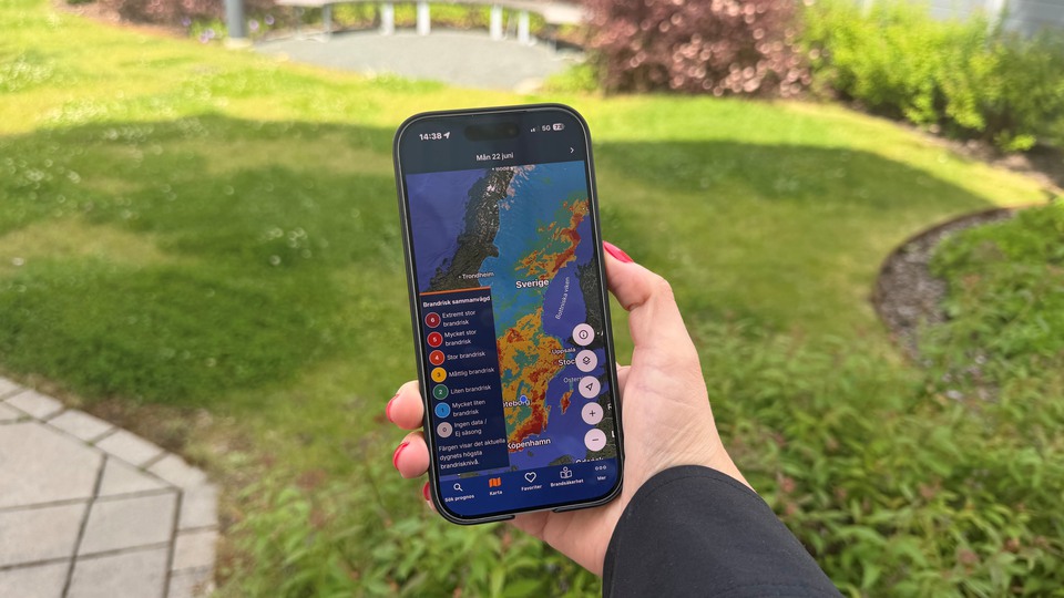 Mobiltelefon visar hur appen Brandrisk ute ser ut. I bakgrunden syns gräsmatta och buskar. Foto.