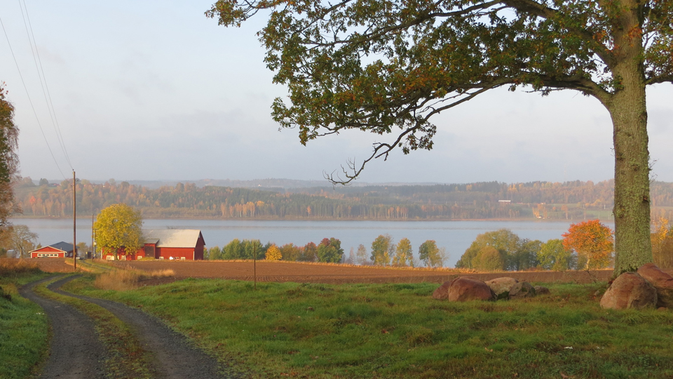 En vy bild över en åker och en sjö med ett litet rött hus vid slutet av vägen.