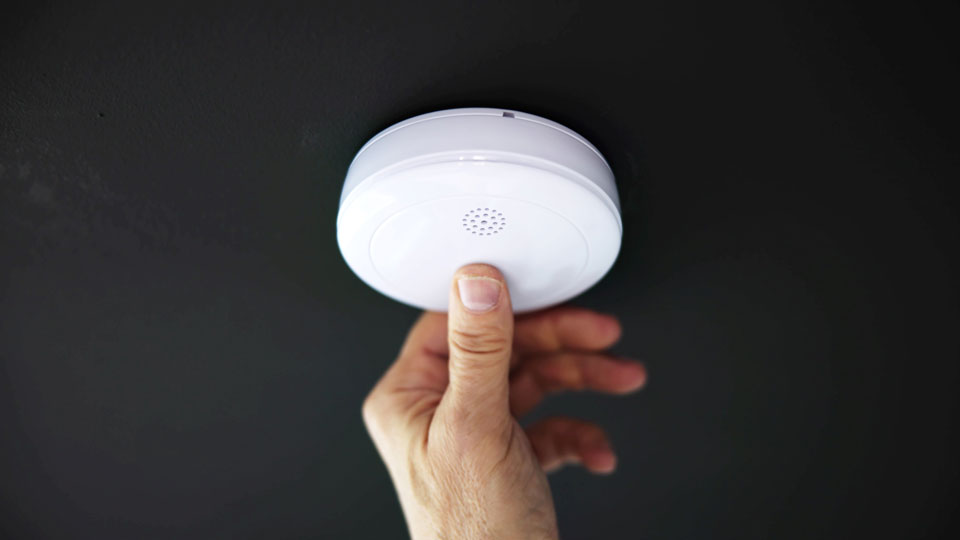 Person trycker på testknappen på en brandvarnare som hänger i taket. Foto.