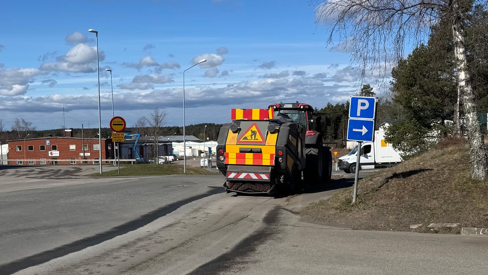 En sopmaskin kör längs en asfalterad väg i ett öppet gatuområde. Maskinen har varningsmarkeringar i rött och gult på baksidan. Till höger syns en blå skylt som visar sväng mot parkering. I bakgrunden finns byggnader, träd och en klar blå himmel med moln.. Foto 