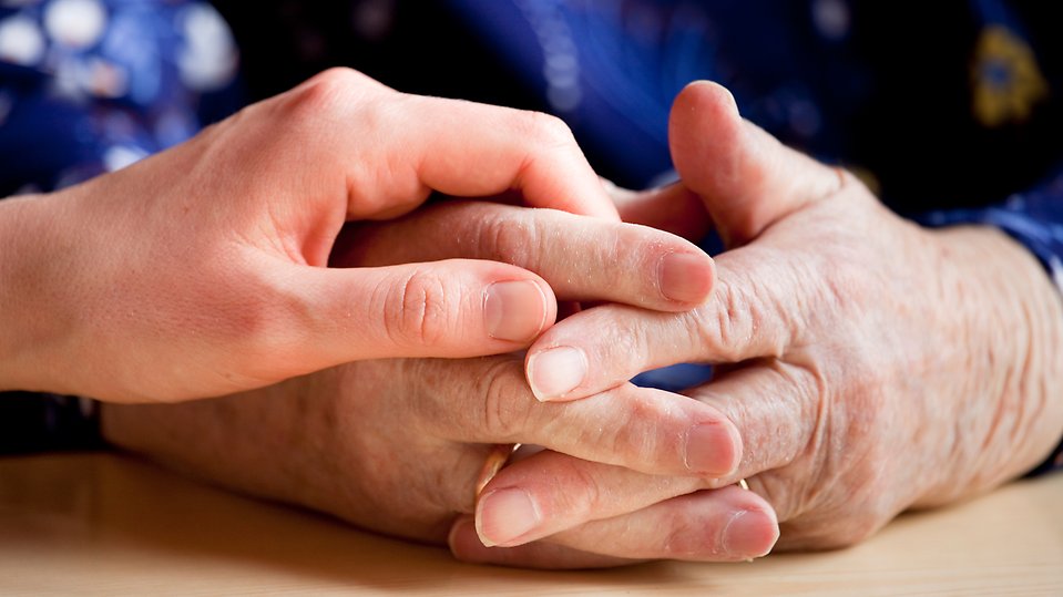 En äldre person som håller ihop sina händer, med en yngre människans hand ovanpå. 