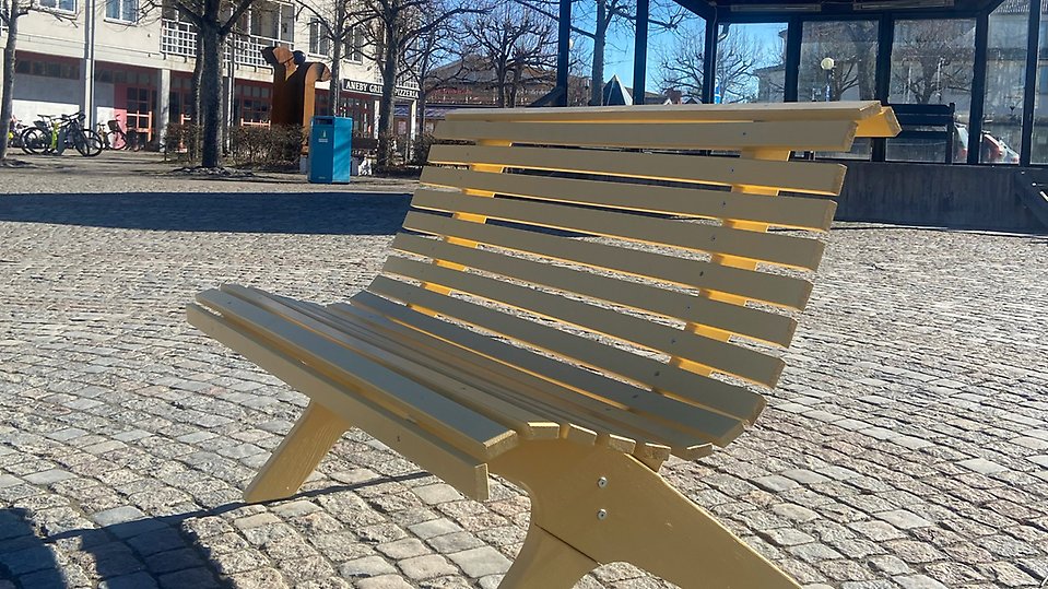 Gul träbänk sedd bakifrån, placerad på en stenlagd öppen yta. Foto