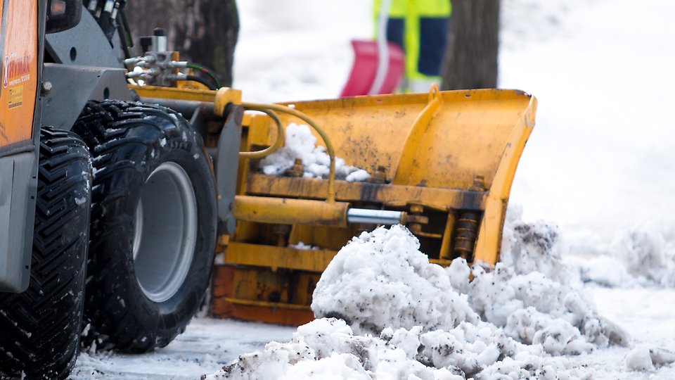 rbild av en snöplog som röjer slaskig snö från en vinterväg. Den gula plogbladet är monterat på ett stort fordon med grova däck, och i bakgrunden syns en person i varselkläder med en röd snöskyffel. Foto 
