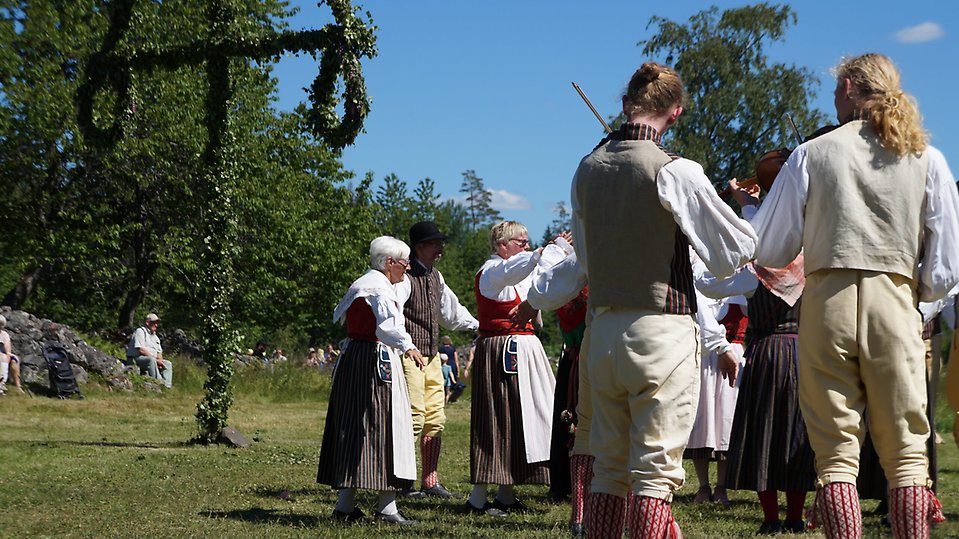 Personer i svenska folkdräkter dansar runt en midsommarstång. Foto. 