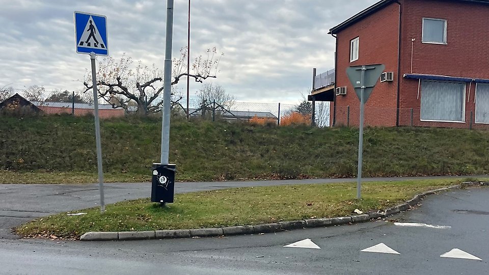 Refug vid vägkorsning med övergångsställesskylt, väjningspliktsskylt och papperskorg, hus i bakgrunden. Foto