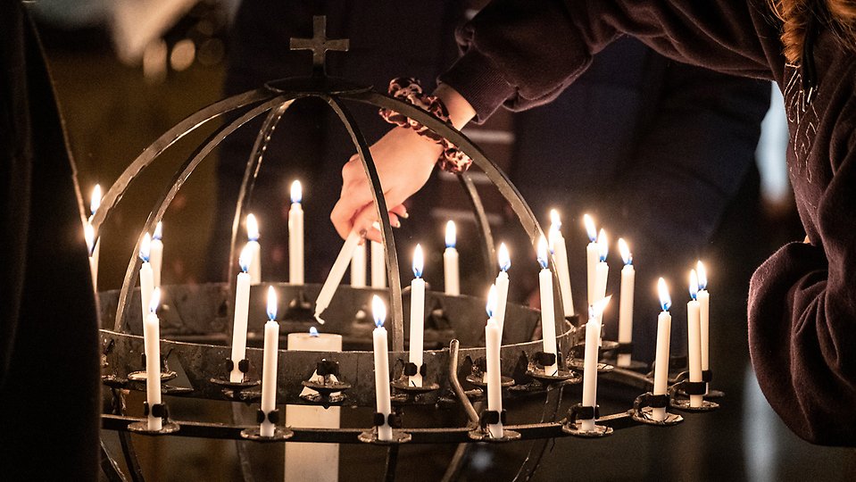 En person tänder ett ljus i en ljushållare fylld med levande ljus i en kyrka. Foto