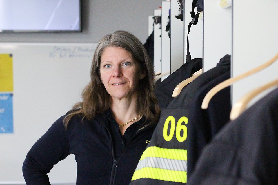 Åsa Karlsson ståendes i rummet där brandmännen byter om. I förgrunden hänger jackor som används ute på olyckor och i bakgrunden står Åsa.