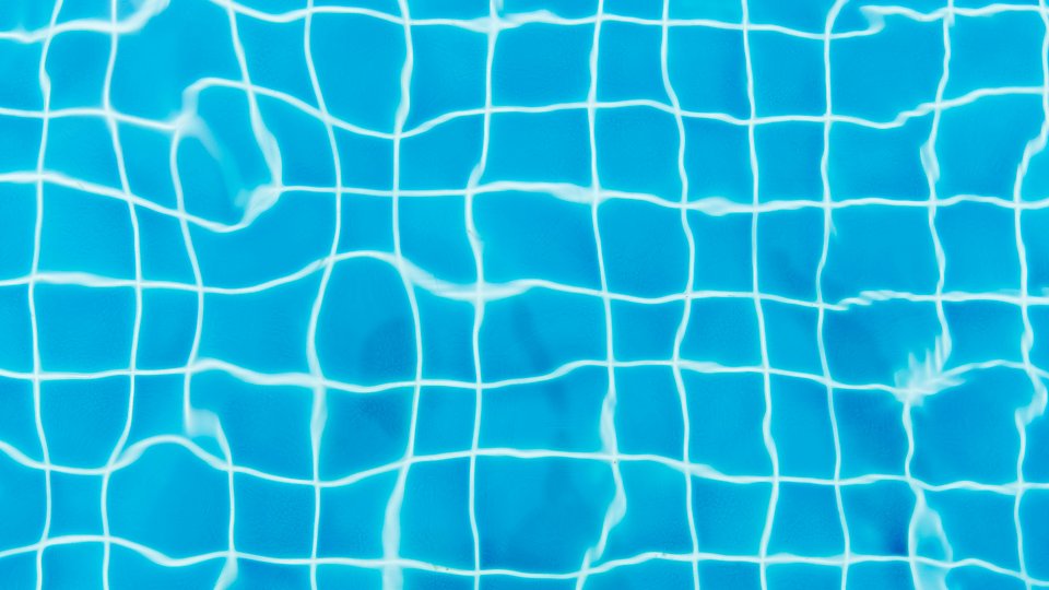 Närbild av ett blått kaklat poolgolv under vatten, med vita linjer som bildar ett rutmönster och ljusreflektioner som skapar vågiga skuggor. Foto